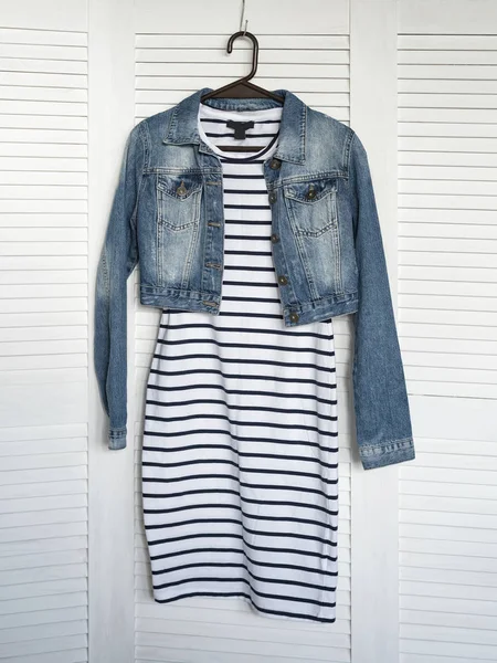 Striped dress and denim jacket on a hanger. White wooden background. Fashionable wardrobe