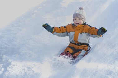 Çocuk şapka ve turuncu tulum içinde sırt üstü kar slaytın dışına kayıyor. Portre. Yakın çekim. Kış günü