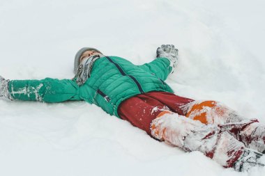 Yeşil bir ceket ve kırmızı pantolon kar sırtına uzanmış silahlarla yatan çocuk. Kış günü