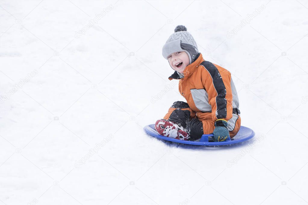 Happy laughing small boy slides down the hill on snow saucer. Seasonal concept. Winter day.