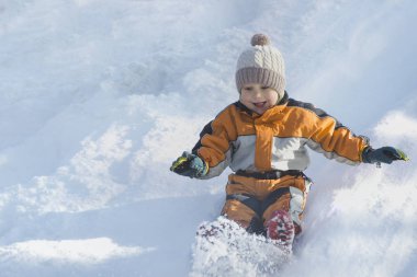 Çocuk şapka ve turuncu tulum içinde sırt üstü kar slaytın dışına kayıyor. Portre. Yakın çekim. Kış günü