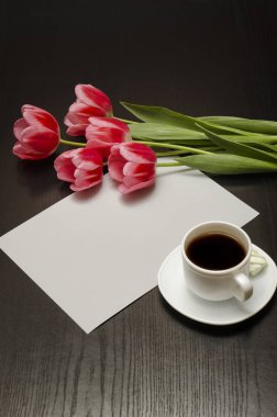 Tatil kavramı. Buket pembe laleler, bir fincan kahve ve siyah bir ahşap zemin üzerine kağıt. Dikey çerçeve.