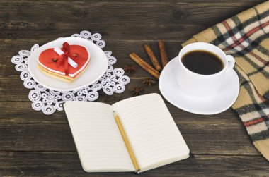 Bir fincan kahve, mesaj, defter, kalem ve kareli ekose kahverengi ahşap bir masa üzerinde kalp şeklinde kurabiye. Tatil planlama
