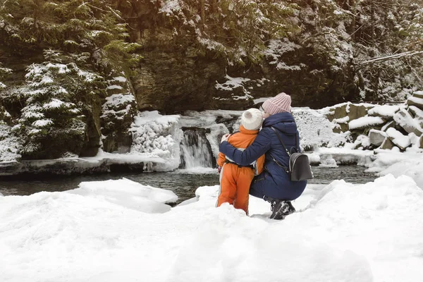 妈妈和儿子坐在白雪覆盖的瀑布 山林和松树林的背景下 拥抱在一起 冬季多云 — 图库照片