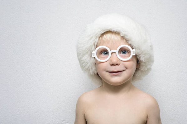 Little boy wearing glasses and a Santa Claus hat stands on the light background. Portrait.
