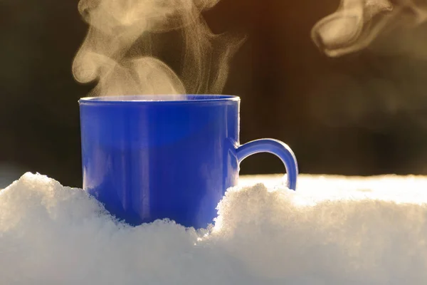 Blue mug with a steaming hot drink is standing in the snow. Winter sunny day.