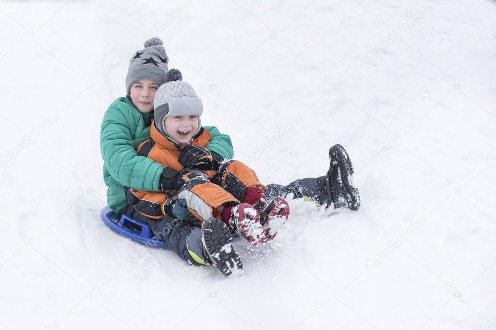 Two joyful boys slides down the hill on snow saucer. Brotherly friendship. Winter day.