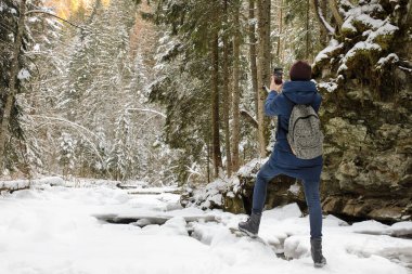Kız bir sırt çantası konumu karla kaplı iğne yapraklı orman içinde olan ve Smartphone'da fotoğraf yapar. Kış günü.
