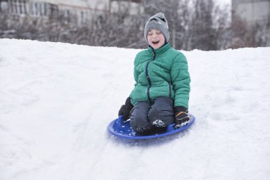 Happy laughing boy with closed eyes slides down the hill on snow saucer. Seasonal concept. Winter day.