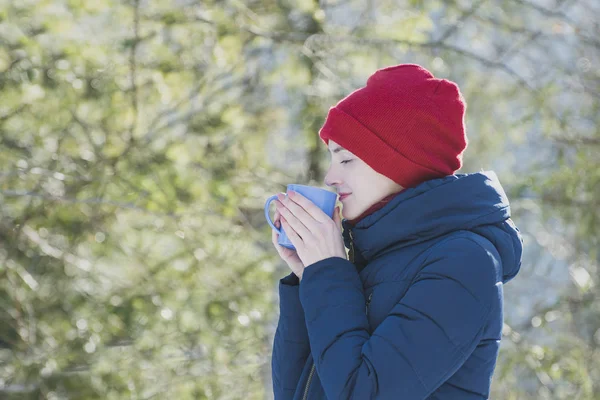 一个闭着眼睛的女孩 戴着一顶红色的帽子 手里拿着一个杯子 站在绿意盎然的背景下 — 图库照片