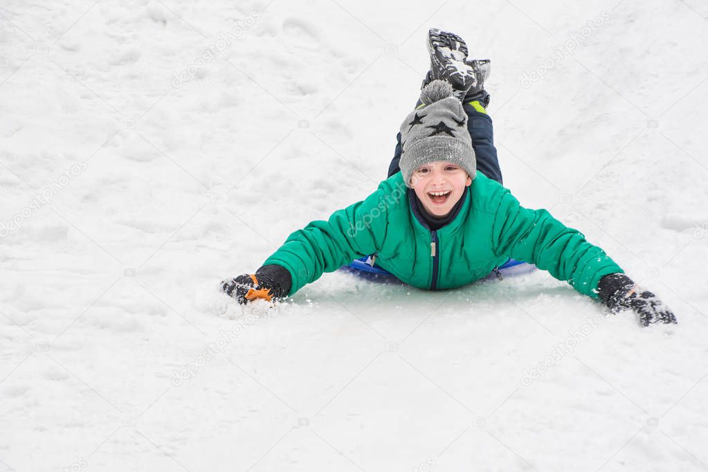 Laughing boy rides on his stomach from a hill on snow saucer. Seasonal concept. Winter day.