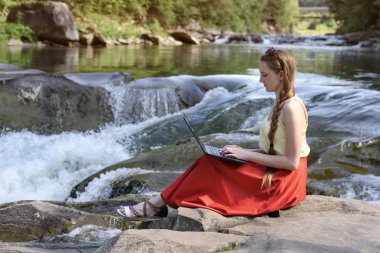 Güzel uzun saçlı kız dağ nehir art arda sıralı bir arka plan üzerinde bir kayaya oturan dizüstü bilgisayar ile kırmızı etek. Serbest kavramı. Doğada çalışma.