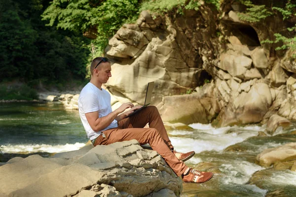 Work in nature concept. Man with notebook sitting at the bank of river over the waterfall and green trees. Side view.