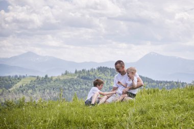 Mutlu baba çim yeşil orman, dağlar ve gökyüzü bulutlu bir arka plan üzerinde oturan iki genç oğlu ile. Dostluk kavramı