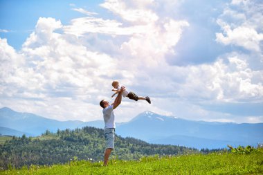 Mutlu baba küçük oğlu yeşil orman, dağlar ve gökyüzü bulutlu bir çerçevede yukarı atar. Babalık dostluk.