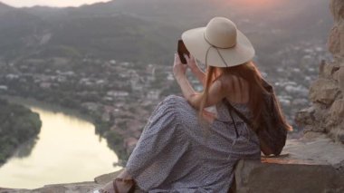 Tepede oturan bir sırt çantası ile şapkalı kız. Smartphone-e doğru şehir aşağıdaki video çekimleri