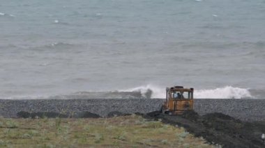 Buldozer fırtınalı denizde zemine karşı kıyı güçlendirmek için çalışıyor. Çoğunlukla bulutlu