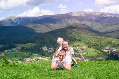 Mutlu bir aile: neşeli baba, anne ve iki oğlu yalan söylüyorsun orman, dağ ve gökyüzü arka planı yeşil çimenlerin üzerinde bulutlar ile.