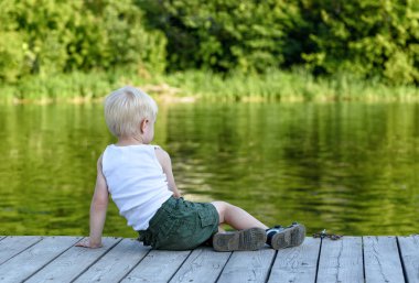 Yeşil bir ormanın ortasında nehir kıyısında bir iskelede oturup küçük sarışın çocuk