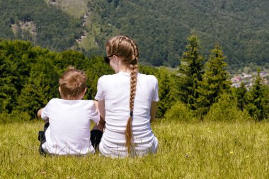 Anne ve iğne yapraklı orman ve dağlar arka plan çimlerde oturan genç oğlu