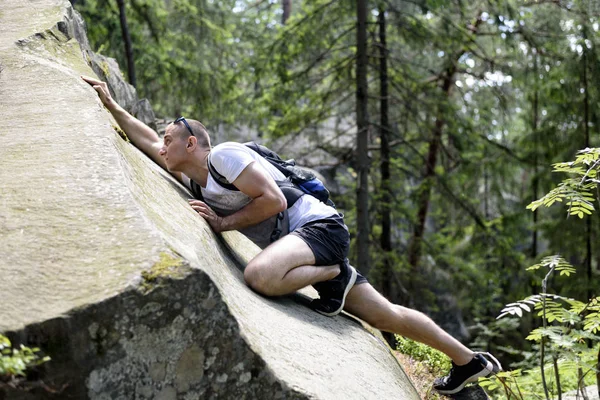 Young man with a backpack climbs up the cliff in the coniferous forest. Close-up — Stock Photo, Image