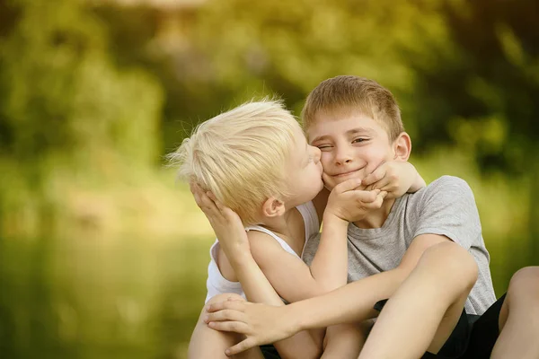 Twee broertjes zitten buiten. Een kussen de andere op de Wang. Wazig groene bomen in de verte. Concept van vriendschap en broederschap. — Stockfoto