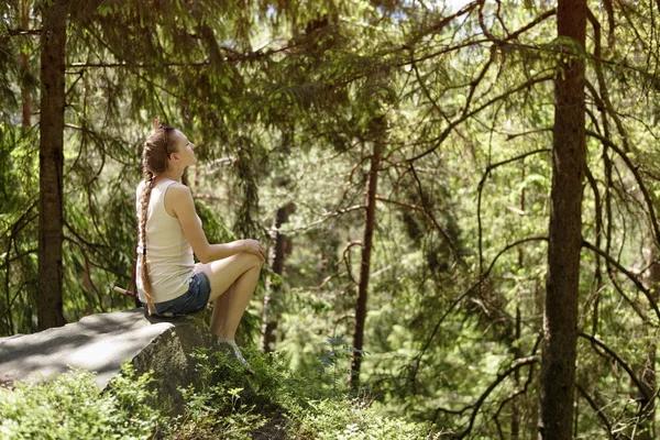 Güneşli bir günde iğne yapraklı orman tarafından çevrili bir taş üzerinde oturan güzel kız rüya