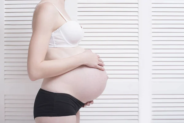 Mujer embarazada en ropa interior abrazando el vientre con las manos sobre un fondo claro . — Foto de Stock