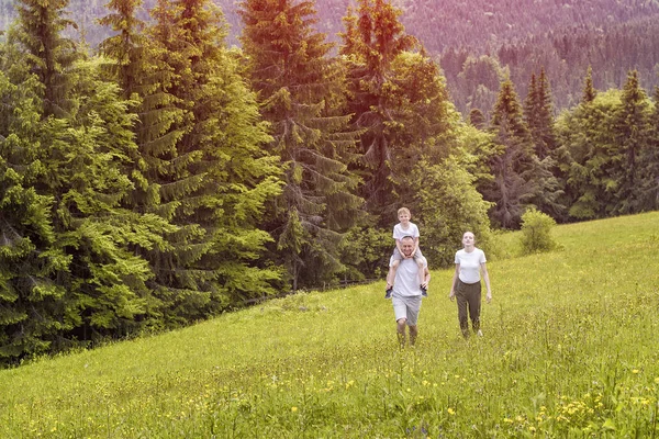 Happy family: father with son on shoulders and mother go on a green field on a background of pine forest — Stock Photo, Image