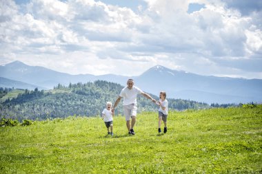 Baba ve yeşil orman, dağlar ve bulutlu gökyüzü arka plan üzerinde el ele tutuşarak yeşil alan çalışan iki genç oğlu.