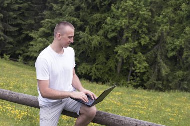 adam ahşap bir çit üzerinde oturur ve bir dizüstü bilgisayar alan ve iğne yapraklı orman ile çalışır