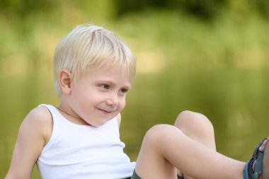 Açık havada bulanık yeşil arka plan üzerinde oturan küçük sarışın çocuk gülümseyen bırakır.