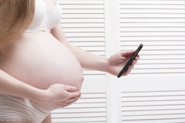 Mujer embarazada en ropa interior cogida de la mano en el estómago y utiliza el teléfono inteligente. Llama a una ambulancia. Contracciones prenatales —  Fotos de Stock