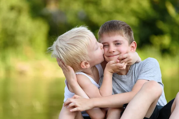 Dos hermanitos están sentados afuera. Uno besa al otro en la mejilla. Árboles verdes borrosos en la distancia. Concepto de amistad y fraternidad . — Foto de Stock