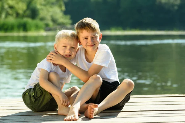 Two little boys sit in an embrace on the banks of the river. Concept of friendship and fraternity — Stock Photo, Image