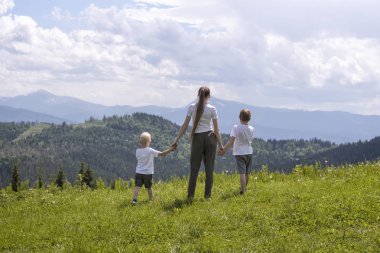 Anne ve iki küçük oğlu tutuşup bir yeşil alan orman, dağlar ve gökyüzü bulutlu bir arka plan üzerinde durmak.