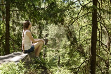 Güneşli bir günde iğne yapraklı orman tarafından çevrili bir taş üzerinde oturan güzel kız rüya