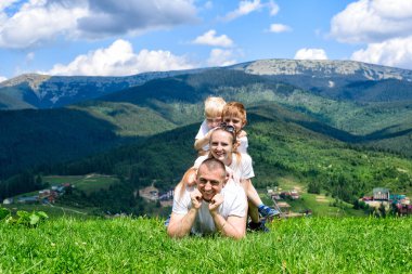 Mutlu bir aile: neşeli baba, anne ve iki oğlu yalan söylüyorsun orman, dağ ve gökyüzü arka planı yeşil çimenlerin üzerinde bulutlar ile.