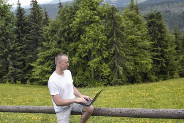 adam ahşap bir çit üzerinde oturur ve bir dizüstü bilgisayar alan ve iğne yapraklı orman ile çalışır
