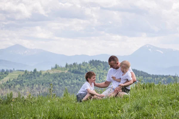 Glücklicher Vater mit seinen beiden kleinen Söhnen, die auf dem Gras vor einem Hintergrund aus grünem Wald, Bergen und wolkenverhangenem Himmel sitzen. Freundschaftskonzept. lizenzfreie Stockfotos