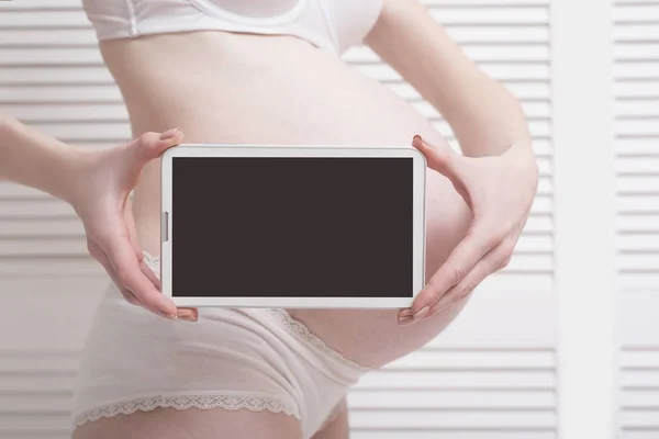 Kobieta w ciąży w bielizny gospodarstwa i Wyświetlono cyfrowego tabletu z pustego ekranu na białym tle drewnianych. Szczelnie-do góry — Zdjęcie stockowe
