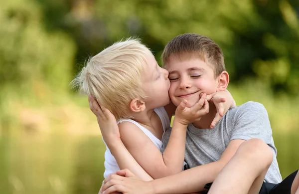 Two little brothers are sitting outdoors. One kisses the other on the cheek. Blurred green trees in the distance. Concept of friendship and fraternity. — Stock Photo, Image