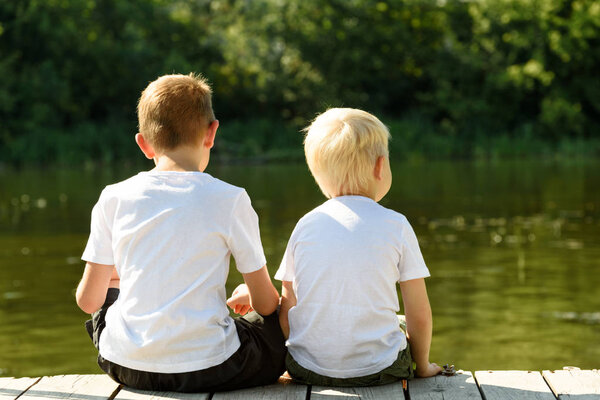 Two little boys are sitting on the pier on the river bank. Concept of friendship and fraternity. Back view