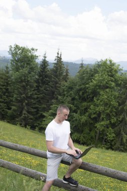 Sürpriz adam ahşap bir çit oturur ve saha ve iğne ağaç orman yakın bir dizüstü bilgisayar arkasında çalışır. Dikey çerçeve