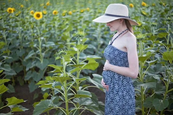 Jeune belle femme enceinte se tient dans un chapeau et une robe sur le terrain de tournesols en fleurs — Photo
