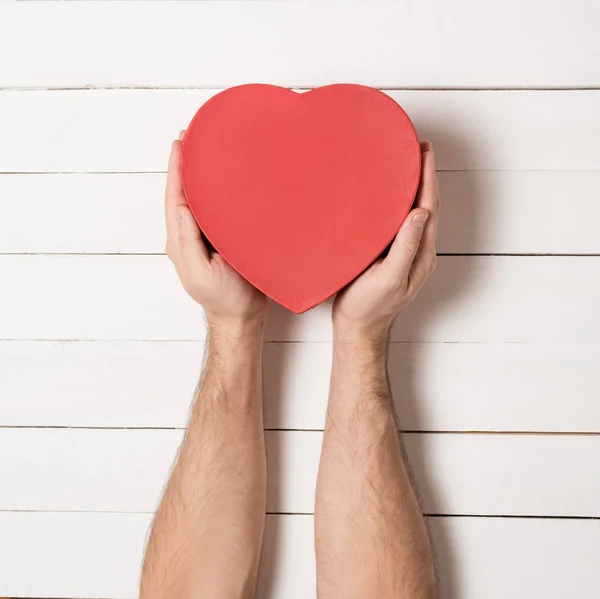 Las manos masculinas sostienen una caja en forma de corazón rojo contra el fondo de una mesa de madera blanca — Foto de Stock