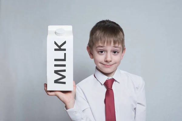 Pequeno menino loiro segura e mostra um pacote de leite grande caixa branca. Camisa branca e gravata vermelha. Fundo de luz — Fotografia de Stock