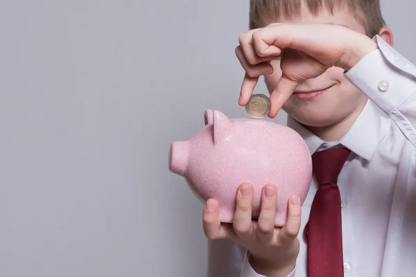 Smiling boy puts a coin in a pink piggy bank. Business concept. Light background