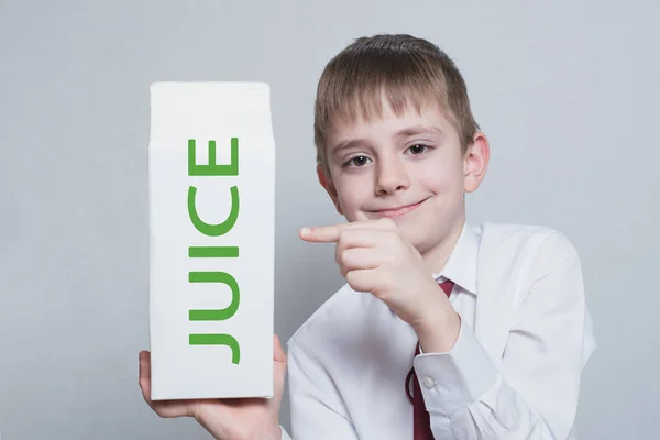 El niño rubio sostiene y muestra el dedo índice en un paquete grande de jugo de cartón blanco. Camisa blanca y corbata roja. Fondo claro — Foto de Stock