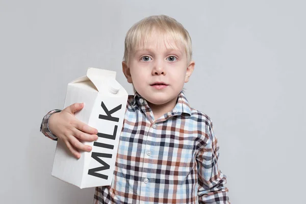 Μικρό ξανθό αγόρι κάνει μια μεγάλη λευκή συσκευασία γάλακτος σε κουτί. Φως φόντο — Φωτογραφία Αρχείου